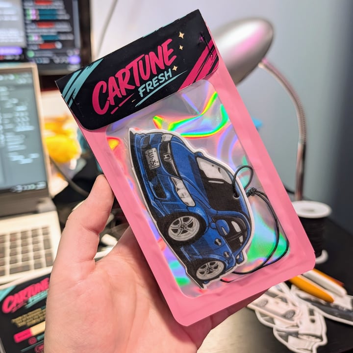 A finished blue car air freshener in pink Cartune Fresh packaging, held in one hand.