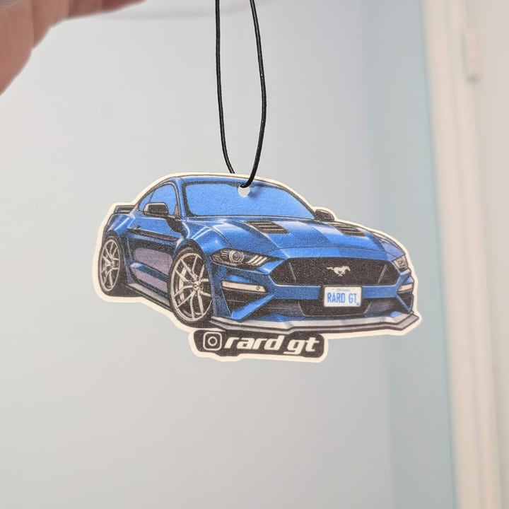 A finished blue sports coupe air freshener hanging from its black elastic loop against a light wall.