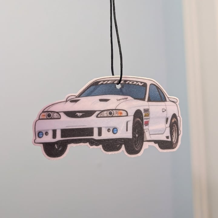 A finished white sports sedan air freshener with a rear wing, hanging against a light wall.